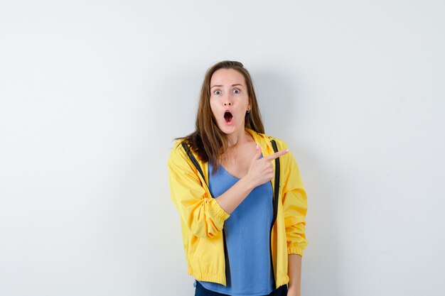 Junge Frau, die auf die obere rechte Ecke in T-Shirt, Jacke zeigt und überrascht aussieht. Vorderansicht.