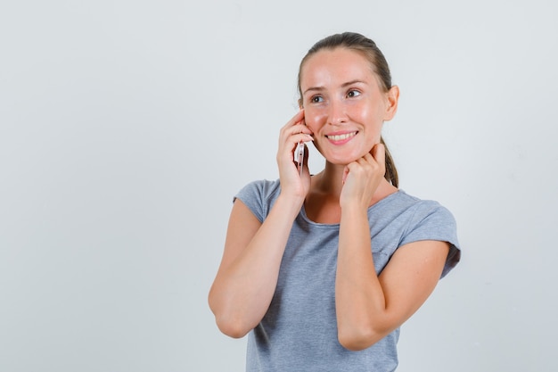 Junge Frau, die auf Handy in grauem T-Shirt spricht und fröhlich schaut. Vorderansicht.