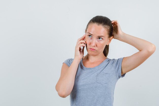 Junge Frau, die auf Handy in grauem T-Shirt spricht und nachdenklich schaut. Vorderansicht.