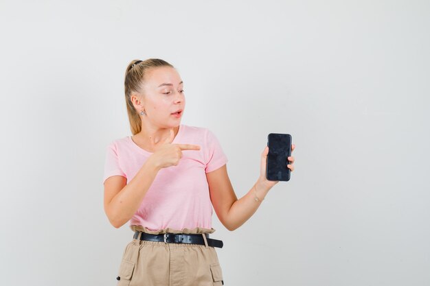 Junge Frau, die auf Handy in T-Shirt, Hosen zeigt und fokussiert, Vorderansicht schaut.