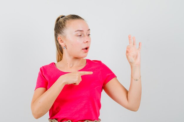 Junge Frau, die auf ihr OK-Zeichen im T-Shirt zeigt und erstaunt schaut