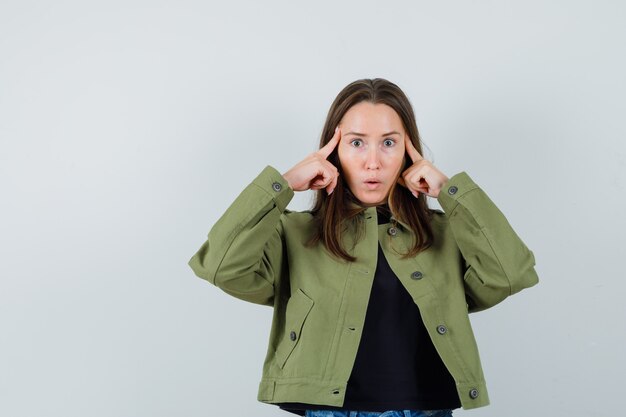 Junge Frau, die auf ihre Schläfe in der grünen Jacke zeigt und konzentriert schaut. Vorderansicht.