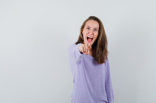 Junge Frau, die auf Kamera zeigt, während sie in lila Bluse schreit und aggressiv schaut. Vorderansicht.