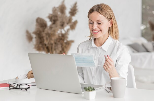 Junge Frau, die auf Laptop während des Haltens einer medizinischen Maske studiert