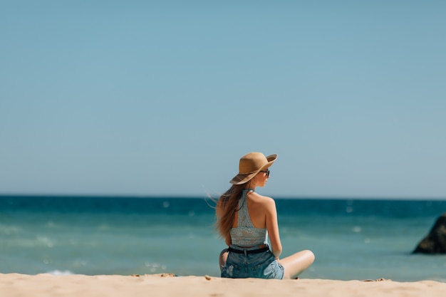 Junge Frau, die auf Sand sitzt und zu einem Meer schaut. Rückansicht