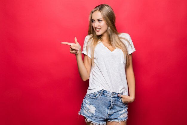 Junge Frau, die auf seitlichen roten Hintergrund zeigt. Sehr frisches und energisches schönes junges Mädchen lächelnd glücklich auf rotem Hintergrund präsentierend.