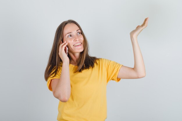 Junge Frau, die auf Smartphone im gelben T-Shirt spricht und fröhlich schaut