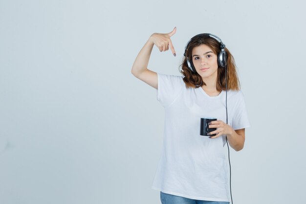 Junge Frau, die auf Tasse des Getränks im weißen T-Shirt zeigt und fröhlich, Vorderansicht schaut.