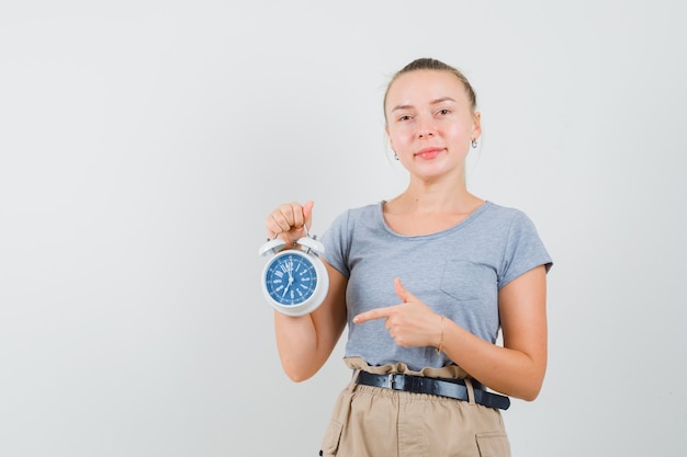 Junge Frau, die auf Wecker im T-Shirt, in den Hosen zeigt und fröhlich schaut. Vorderansicht.