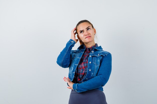 Junge Frau, die aufschaut, während sie den Kopf in kariertem Hemd, Jacke, Hose kratzt und nachdenklich aussieht. Vorderansicht.