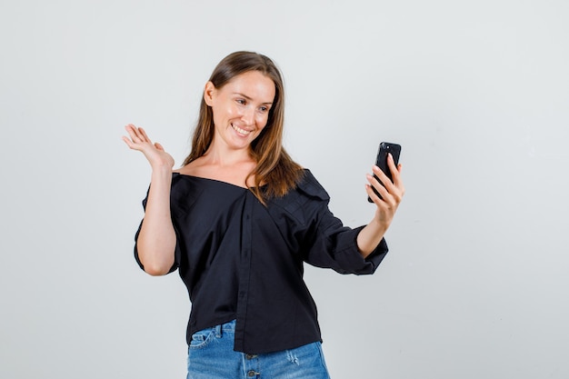 Junge Frau, die aufwirft, während sie selfie auf Smartphone in Hemd, Shorts nimmt und niedlich schaut. Vorderansicht.