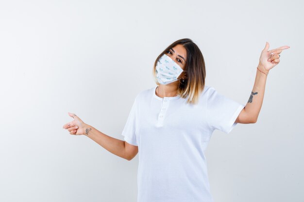 Junge Frau, die beide Seiten im T-Shirt, in der Maske zeigt und fröhlich schaut, Vorderansicht.