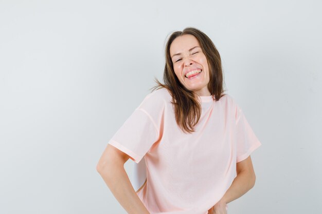 Junge Frau, die beim Augenzwinkern aufwirft, Zunge in rosa T-Shirt herausstreckt und glücklich schaut. Vorderansicht.