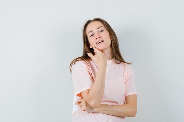 Junge Frau, die beim Stehen im rosa T-Shirt aufwirft und verführerisch schaut. Vorderansicht.