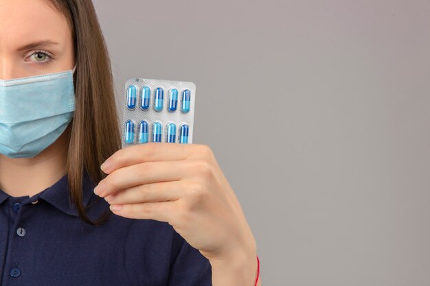 Junge Frau, die blaues Poloshirt in der medizinischen Maske trägt, die ernsthaft Kamera betrachtet, die Blister mit Pillen in der Hand auf hellgrauem lokalisiertem Hintergrund hält
