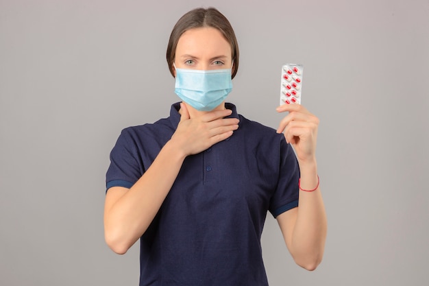 Junge Frau, die blaues Poloshirt in der medizinischen Schutzmaske hält, die Blisterpillen in der Hand hält, die ihren Hals stehend auf hellgrauem Hintergrund berührt