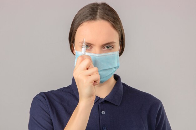 Junge Frau, die blaues Poloshirt in der medizinischen Schutzmaske hält, die eine Ampulle mit einem Impfstoff betrachtet, der Kamera mit ernstem Gesicht betrachtet, das auf lokalisiertem grauem Hintergrund steht