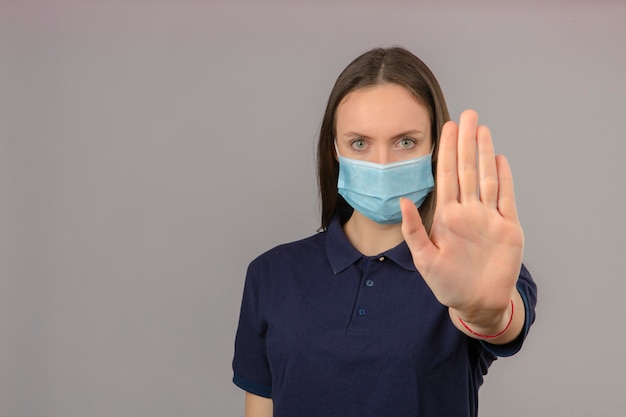 Junge Frau, die blaues Poloshirt in der medizinischen Schutzmaske trägt, die Handstoppgeste mit ernstem Gesicht lokalisiert auf hellgrauem Hintergrund mit Kopienraum zeigt