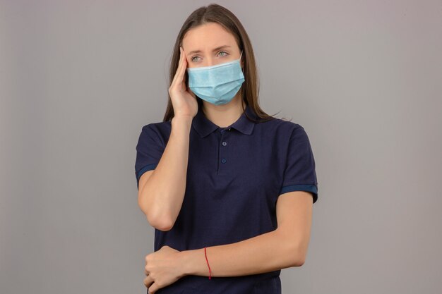Junge Frau, die blaues Poloshirt in der medizinischen Schutzmaske trägt, die Kopfgefühlsschmerz lokalisiert auf hellgrauem Hintergrund berührt