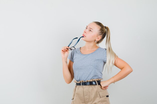 Junge Frau, die Brille hält, während sie in T-Shirt, Hose und nachdenklich schaut. Vorderansicht.