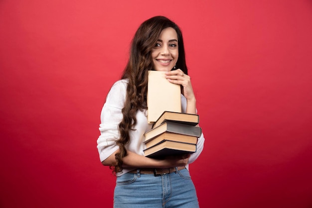 Junge Frau, die Bücher trägt und auf einem roten Hintergrund lächelt. Foto in hoher Qualität