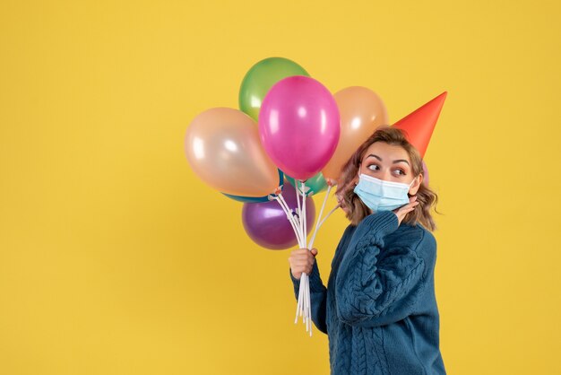 junge Frau, die bunte Luftballons in der Maske auf Gelb hält