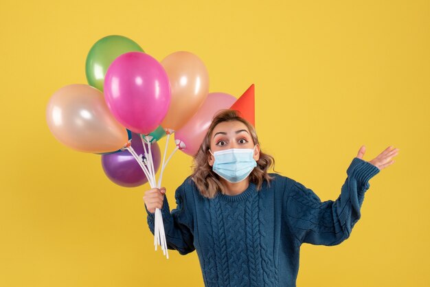 junge Frau, die bunte Luftballons in der Maske auf Gelb hält