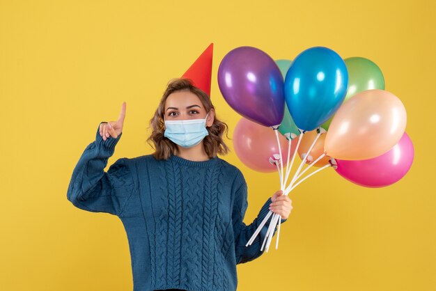 junge Frau, die bunte Luftballons in der Maske auf Gelb hält