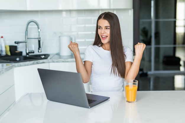 Junge frau, die computer-laptop an der küche verwendet, die stolz schreit und sieg und erfolg sehr aufgeregt feiert