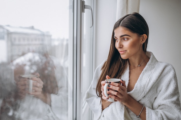 Junge Frau, die das Fenster trinkt heißen Kaffee bereitsteht