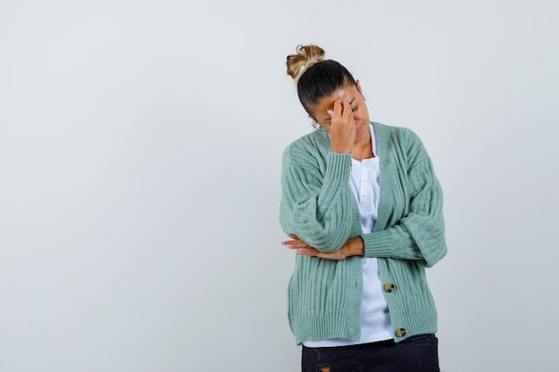 Kostenloses Foto junge frau, die das gesicht mit der hand bedeckt, während sie die hand am ellbogen in weißem hemd und mintgrüner strickjacke hält und gehetzt aussieht