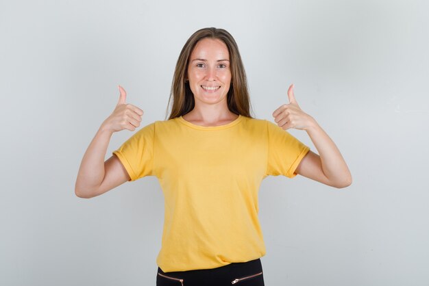Junge Frau, die Daumen oben im gelben T-Shirt, in der Hose und im fröhlichen Blick zeigt
