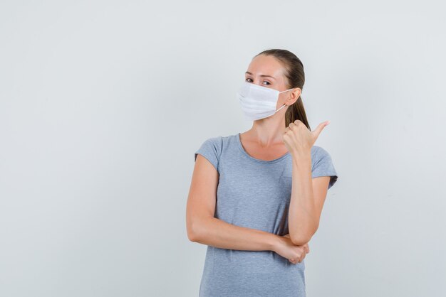 Junge Frau, die Daumen oben im grauen T-Shirt, Maskenvoransicht zeigt.