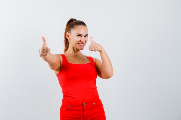 Junge Frau, die Daumen oben im roten Trägershirt, in den Hosen und in der glücklichen Vorderansicht zeigt.