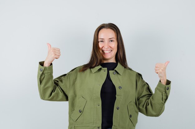 Junge Frau, die Daumen oben in der grünen Jacke zeigt und positiv schaut. Vorderansicht.