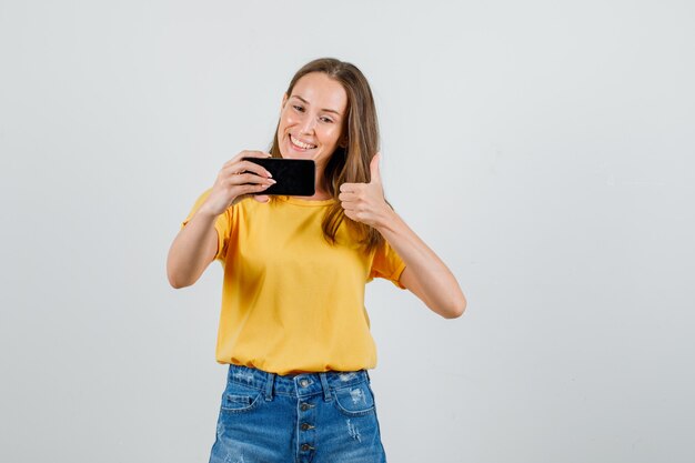 Junge Frau, die Daumen zeigt, während Selfie in T-Shirt, Shorts nimmt und froh aussieht. Vorderansicht.