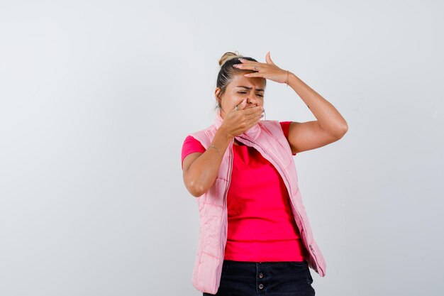 Junge Frau, die den Mund mit der Hand bedeckt, eine Hand auf die Stirn in rosa T-Shirt und Jacke legt und traurig aussieht