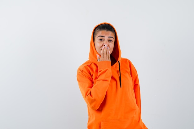 Junge Frau, die den Mund mit der Hand im orangefarbenen Hoodie bedeckt und glücklich schaut