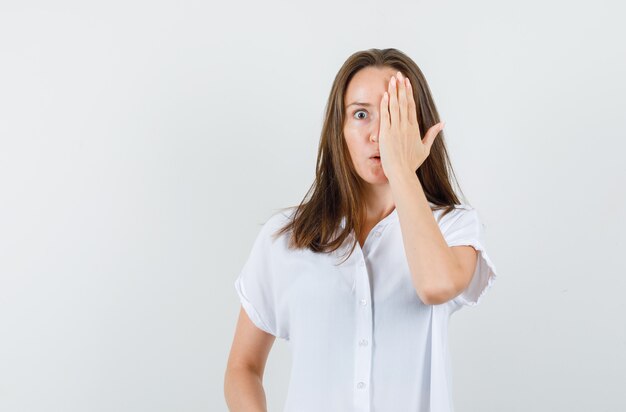 Junge Frau, die die Hälfte ihres Gesichts mit der Hand in der weißen Bluse bedeckt und seltsam aussieht