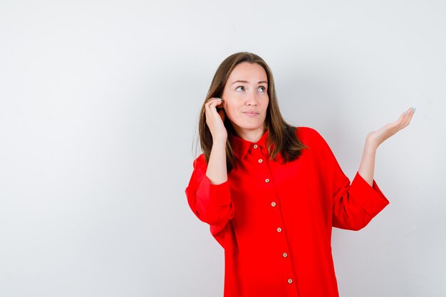Junge Frau, die die Hand in der Nähe des Ohrs hält, die Handfläche in der roten Bluse beiseite ausbreitet und fokussiert aussieht. Vorderansicht.