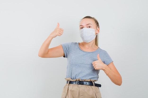 Junge Frau, die doppelte Daumen oben im T-Shirt, in der Hose, in der Vorderansicht der Maske zeigt.