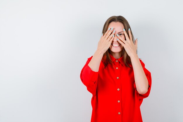 Junge Frau, die durch die Finger in der roten Bluse schaut und fröhlich aussieht, Vorderansicht.