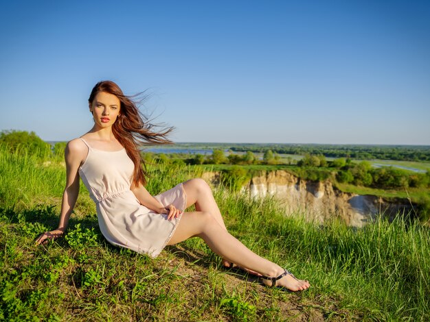 Junge Frau, die durch eine Klippe draußen auf Natur sitzt. Attraktives Mädchen mit einem weißen Kleid, das im Freien aufwirft. Weibliches Modell, das in einem Feld einen sonnigen Sommertag aufwirft.