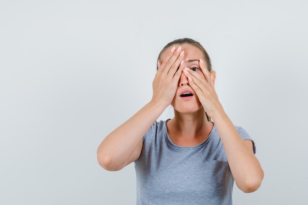 Junge Frau, die durch Finger mit Hand auf Auge im grauen T-Shirt, Vorderansicht schaut.