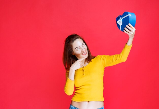 Junge Frau, die eine blaue Geschenkbox in Herzform hält und sich positiv und glücklich fühlt