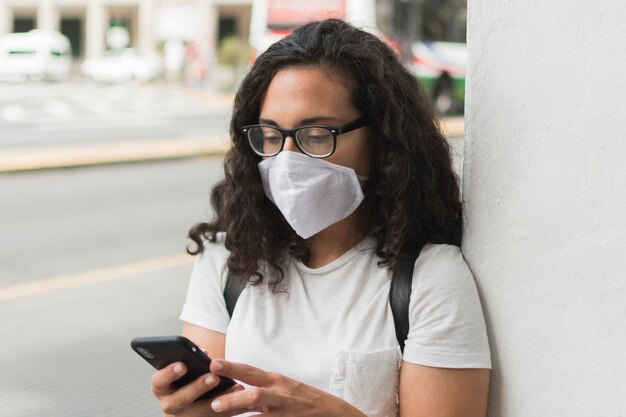 Junge Frau, die eine medizinische Maske trägt, während sie ihr Telefon überprüft