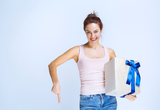 Junge Frau, die eine mit blauem Band umwickelte weiße Geschenkbox hält und die Person neben ihr einlädt, sie zu präsentieren