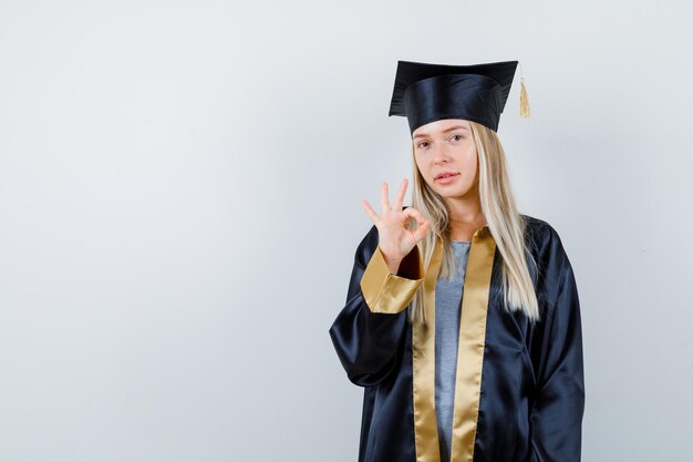 Junge Frau, die eine ok Geste in der Absolventenuniform zeigt und selbstbewusst aussieht.