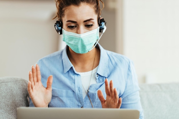 Junge Frau, die eine schützende Gesichtsmaske trägt, während sie den Computer benutzt und zu Hause einen Videoanruf führt