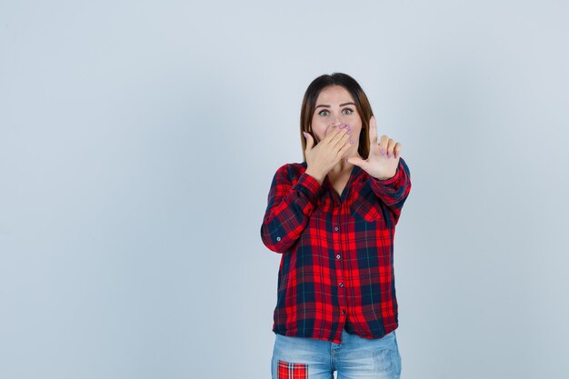 Junge Frau, die eine winzige Geste hält, den Mund mit der Hand in einem karierten Hemd bedeckt und überrascht aussieht. Vorderansicht.